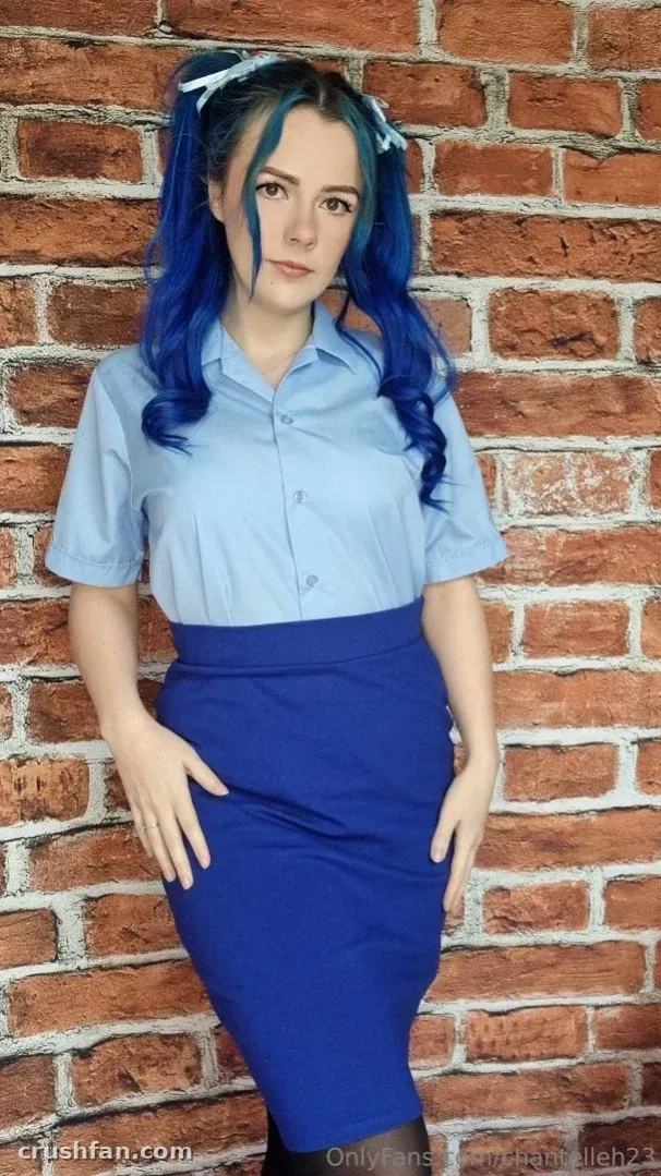 Chantelleh23 dans une chemise bleue et une jupe bleu marine debout près d'un mur de briques.