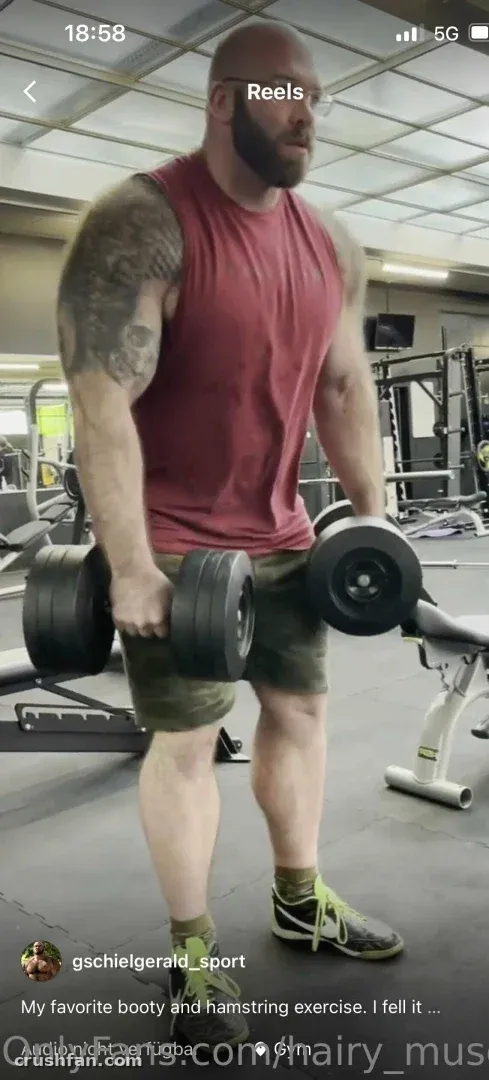 Gschiel Gerald sosteniendo mancuernas en un gimnasio, llevando una camiseta roja sin mangas y pantalones cortos de camuflaje.