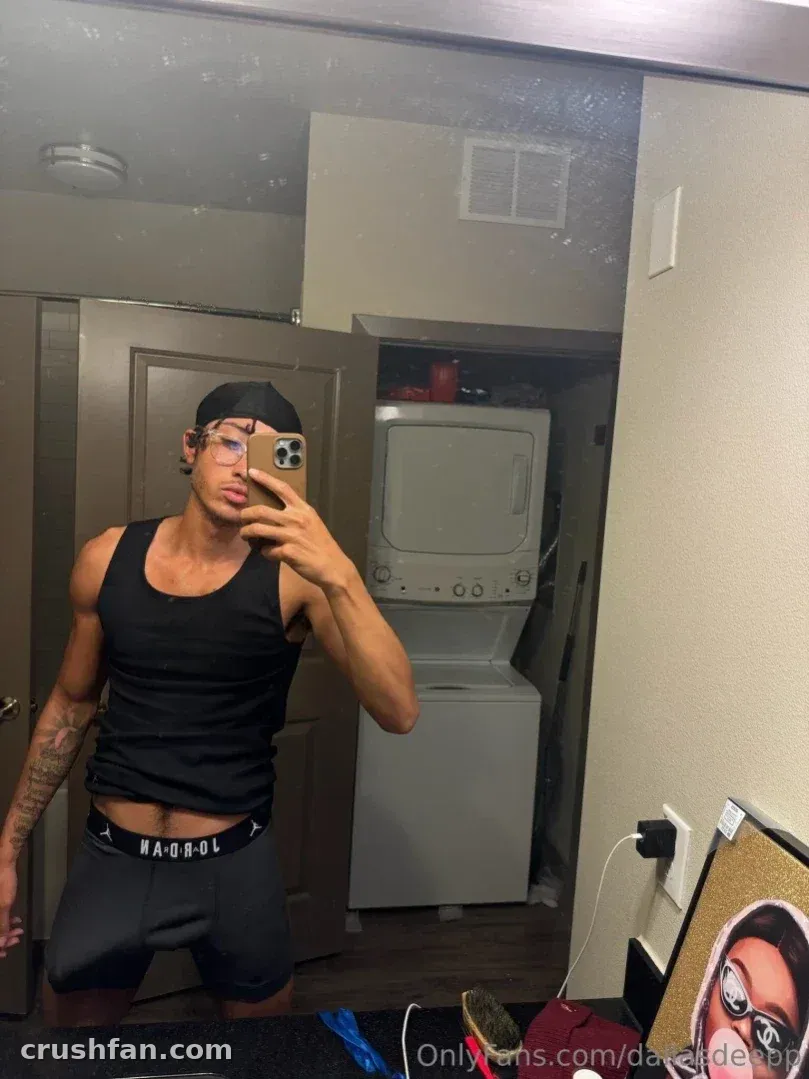 Dallas taking a mirror selfie in a laundry room wearing a black tank top and cap.