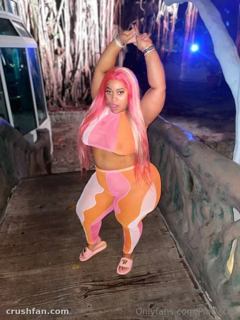 The Official Pinky XXX in an orange and pink two piece outfit standing on a wooden walkway at night.
