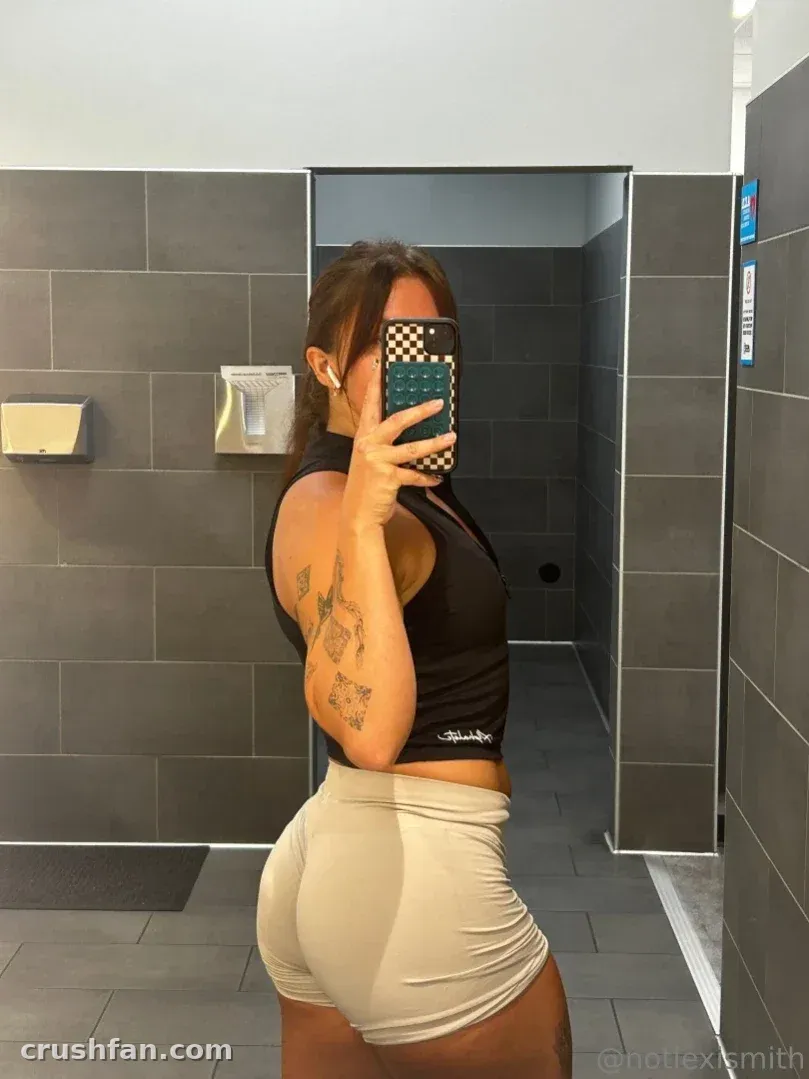Lexi macht ein Spiegel-Selfie in einem schwarzen Crop-Top und beiger Shorts in einem gefliesten Raum.