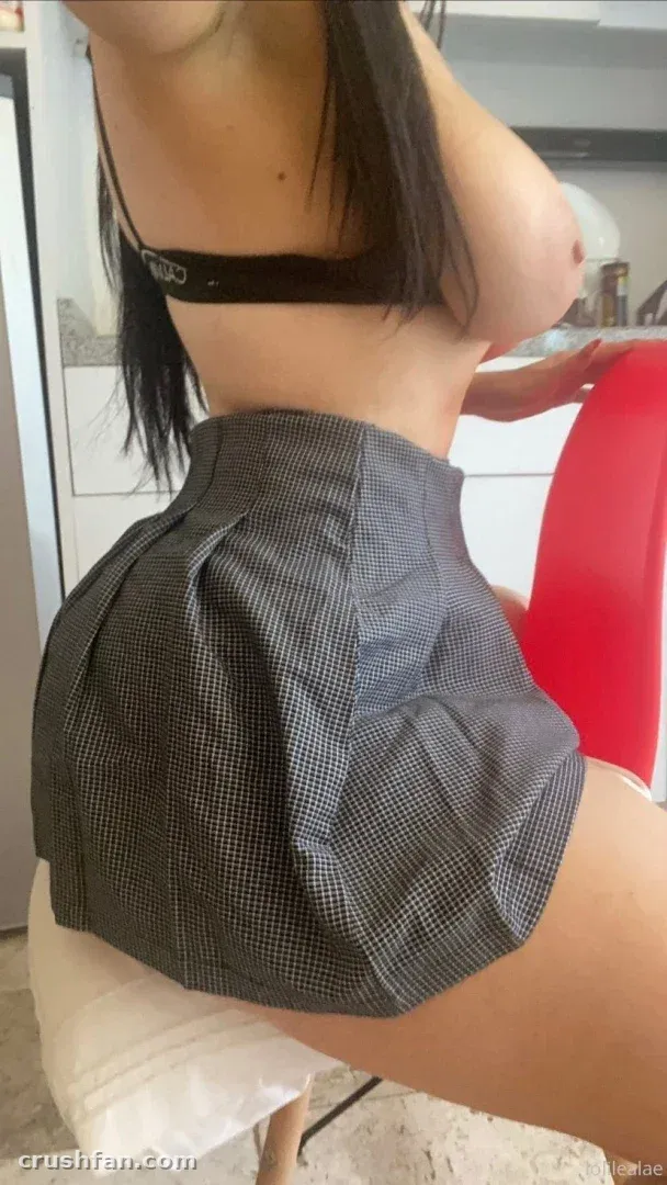 Creator in a black bra and grey high-waisted skirt standing indoors beside a red chair