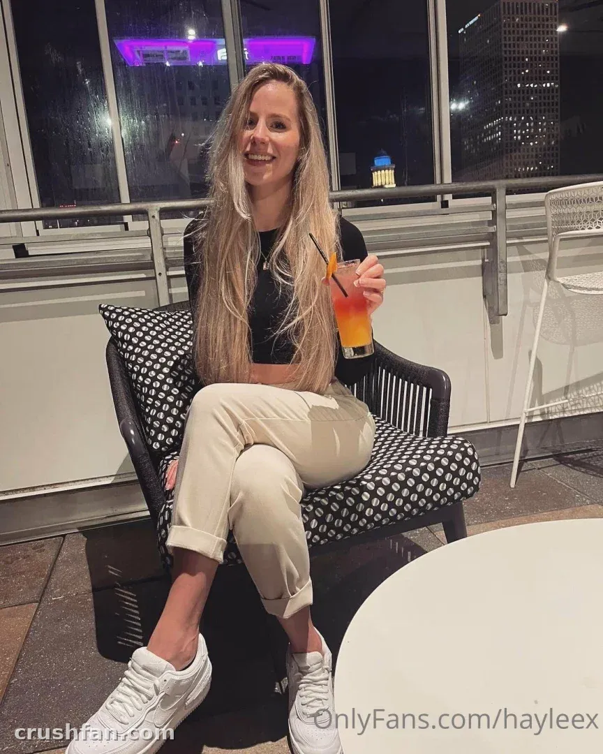 Hayleex assise sur un balcon la nuit tenant un cocktail, portant un haut noir et un pantalon beige.