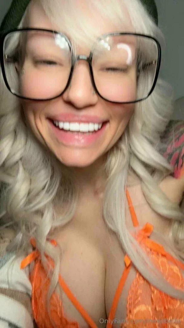Smiling woman with platinum blonde hair, large glasses, green beanie and orange lace top in a close-up selfie.