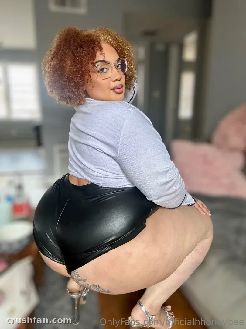 Honey com um top cinza e shorts pretos posando em ambiente interno com cabelo cacheado e óculos