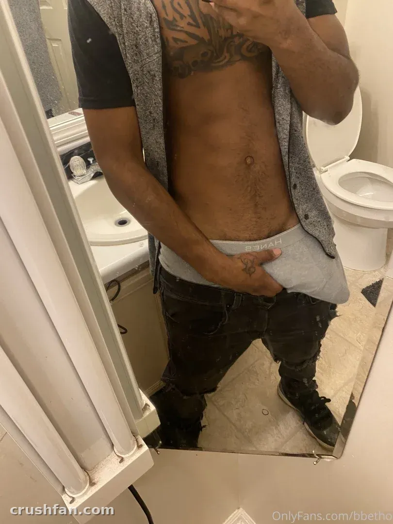 SHE FREAK ETO tomándose una selfie en el espejo de un baño con un chaleco gris y jeans oscuros.