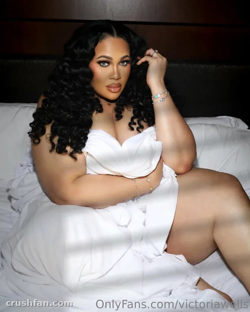 Victoria Wells in a white dress seated on white bedding against a dark headboard