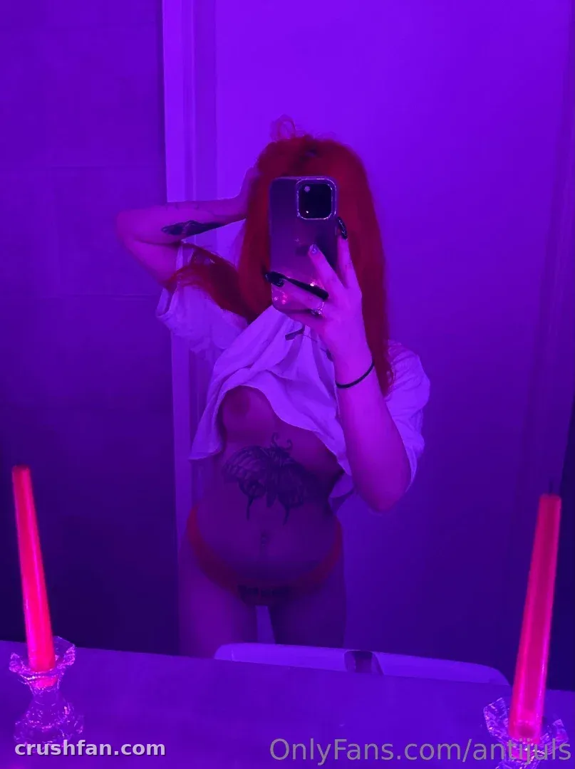 Selfie no espelho de uma mulher de cabelo ruivo sob luz violeta com velas rosa em primeiro plano