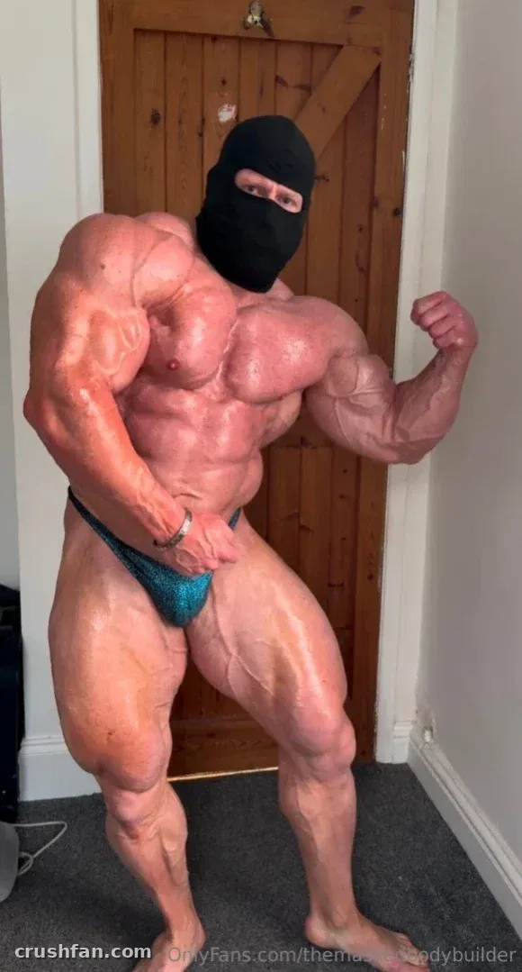 Muscular masked bodybuilder in a teal posing suit flexing indoors beside a wooden door