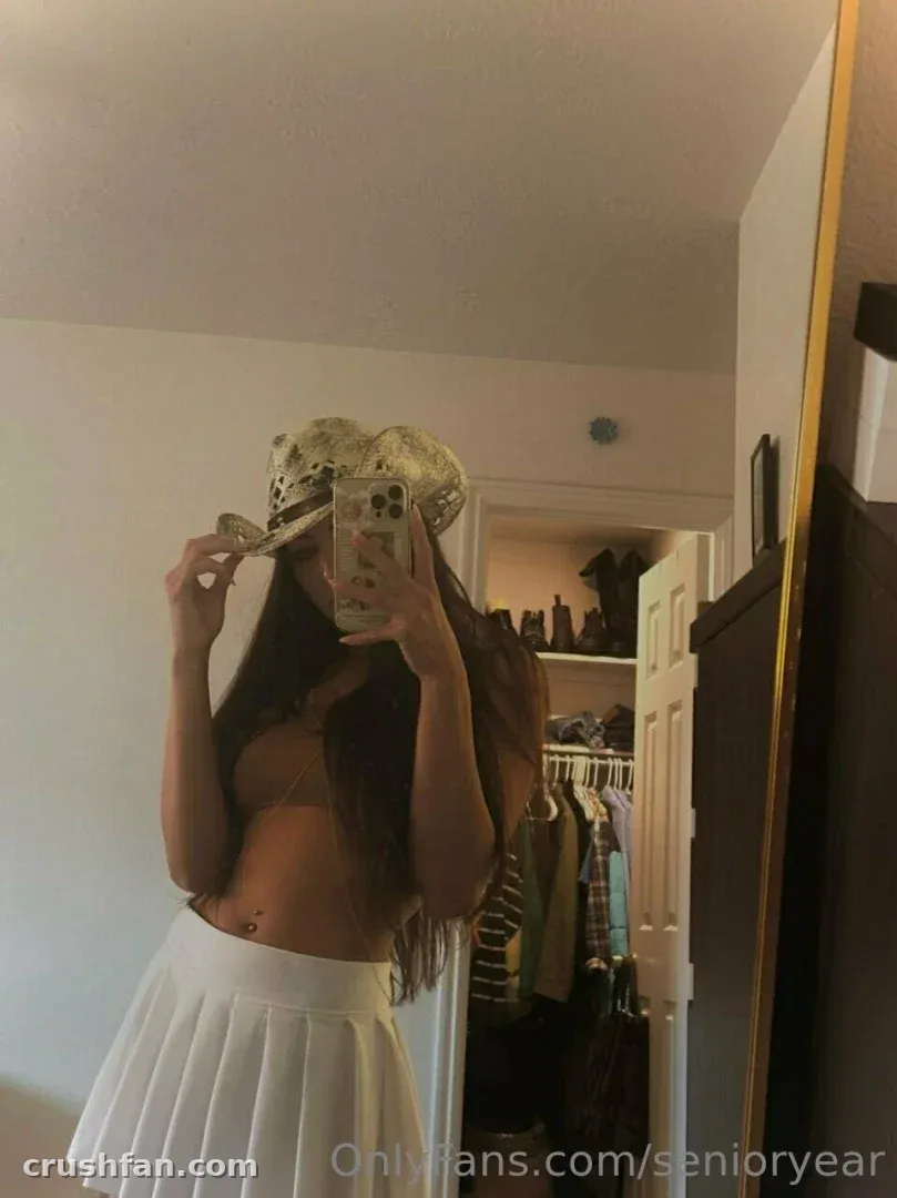 Alexis usando chapéu de renda e saia plissada branca em uma selfie no espelho em ambiente interno