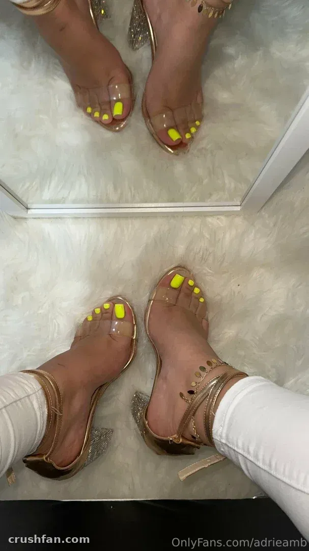 Adripiecitos olhando para baixo, para os pés, em saltos transparentes com unhas amarelas sobre um tapete branco.
