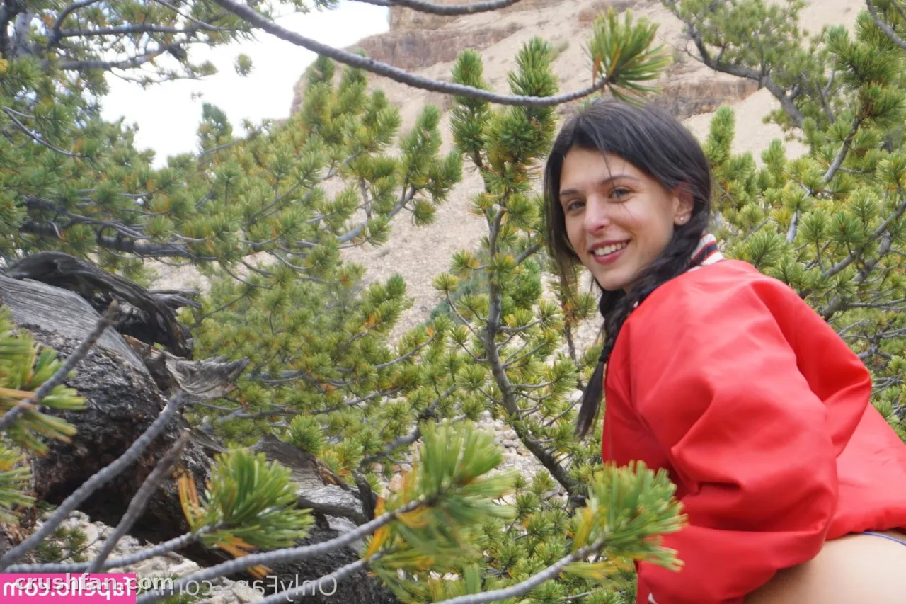 Mulher sorridente de jaqueta vermelha entre galhos de pinheiro em uma encosta rochosa