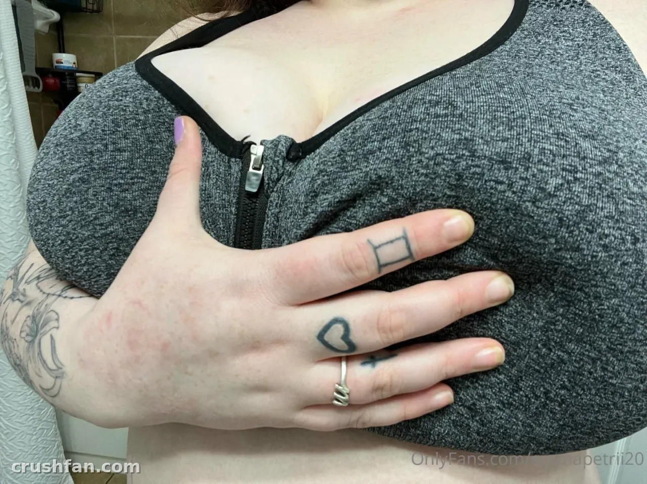 Creadora con un top gris con cremallera delantera, tatuajes y un anillo de plata, fotografiada en interiores