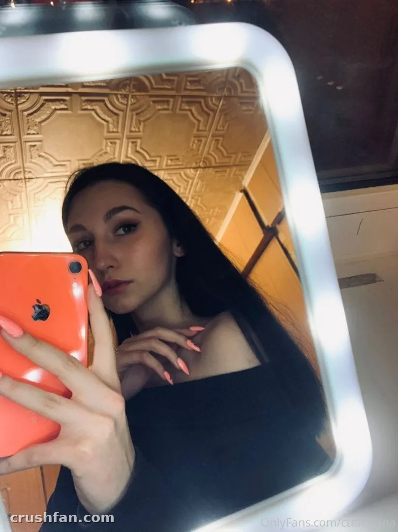 Cutie Lana делает селфи в зеркале с оранжевым телефоном в комнате с тёплыми тонами.