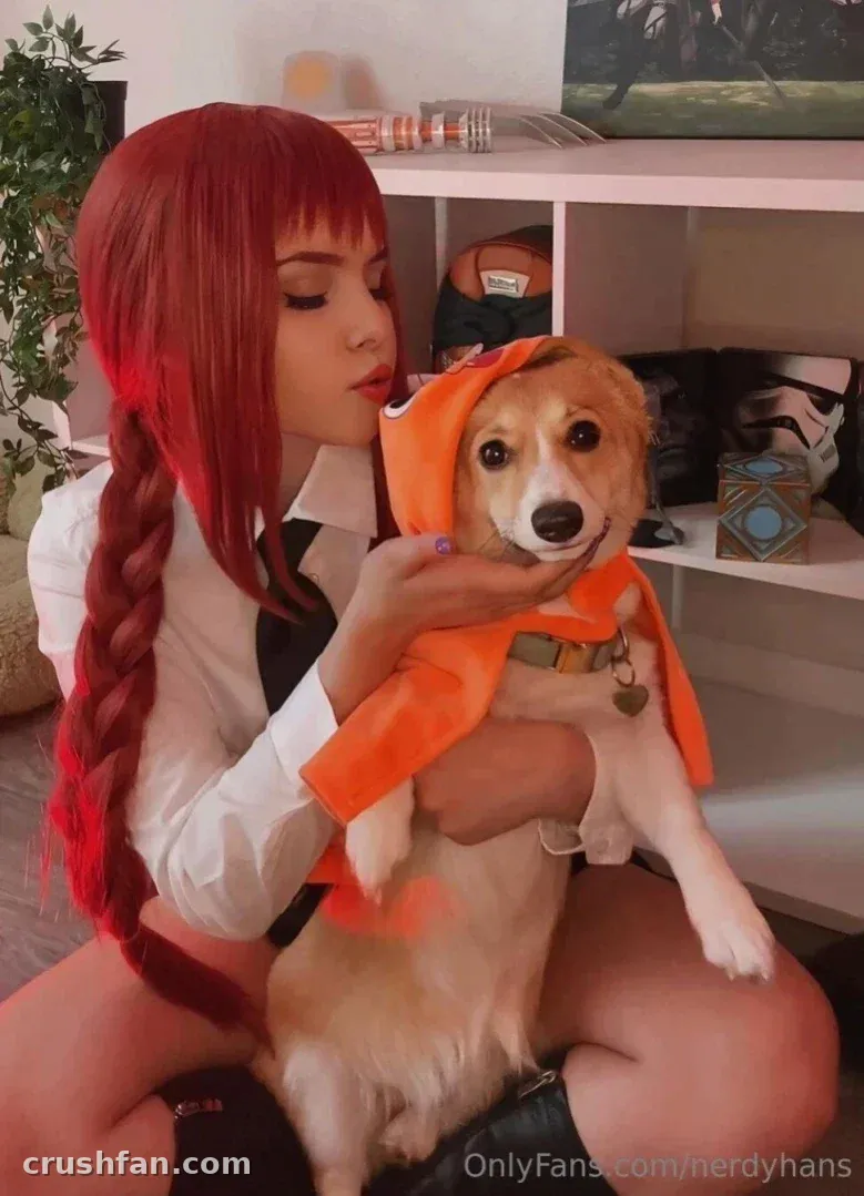 Creator with a long red braid holds a small dog wearing an orange hooded cape indoors.
