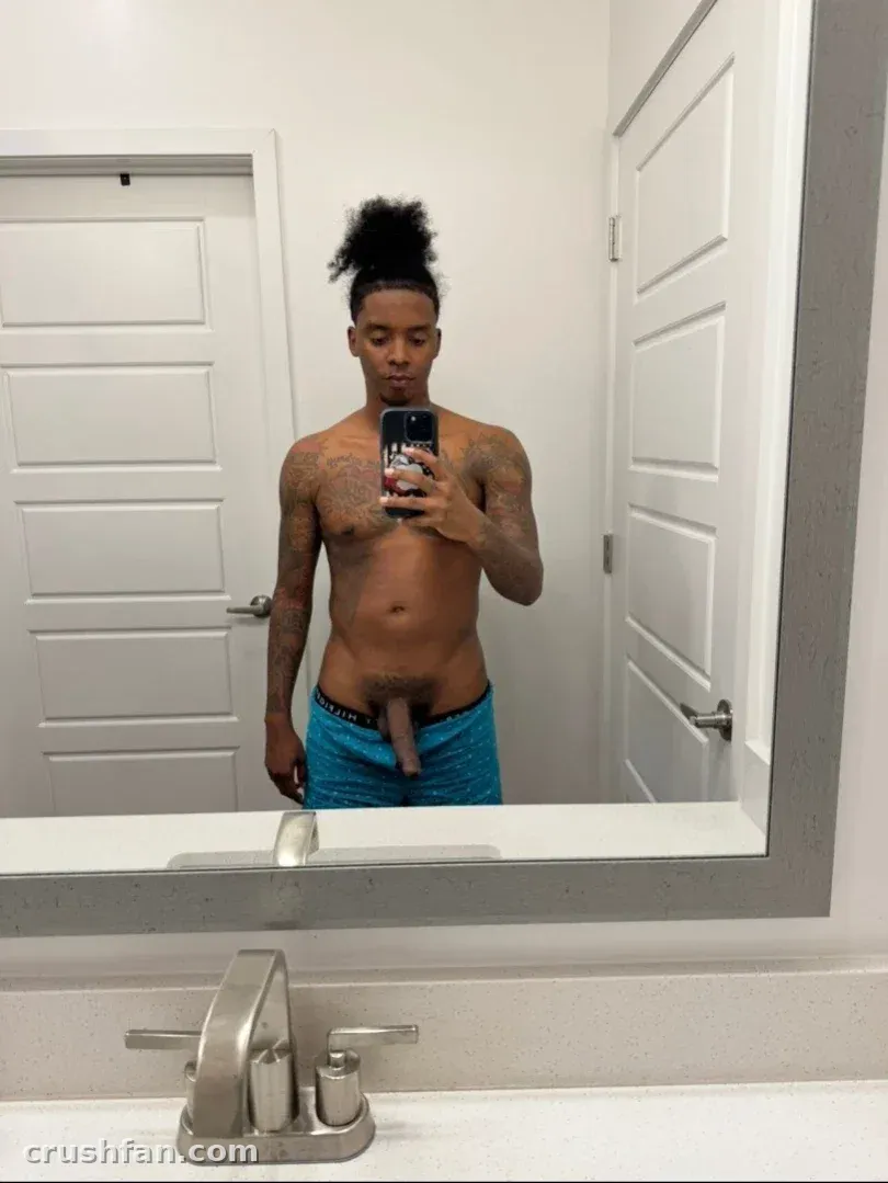 Shirtless creator with tattoos and a topknot takes a mirror selfie in a bathroom.