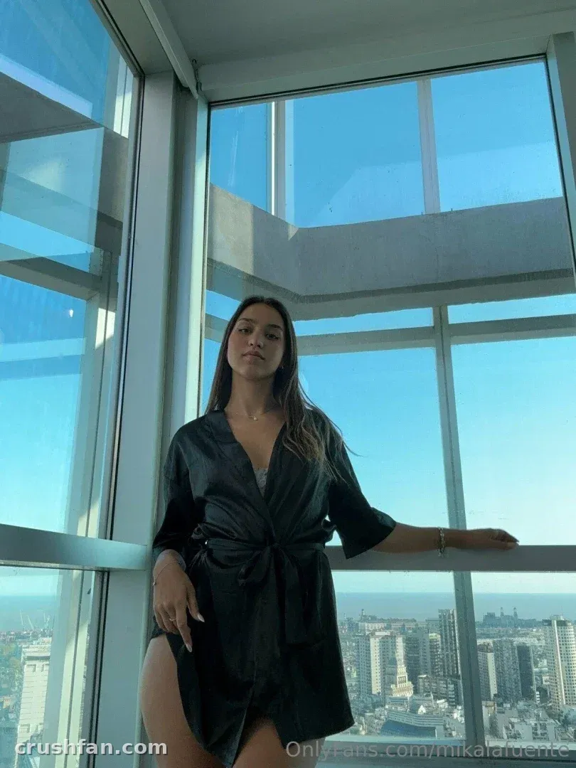 Mika in a black satin robe standing by a high-rise window with a city skyline behind her