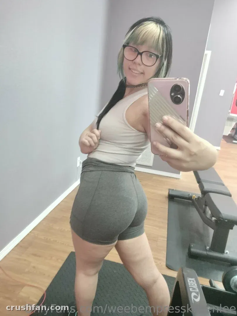 EmpressKoneko se toma un selfie frente al espejo en un gimnasio con una camiseta sin mangas blanca y shorts grises.
