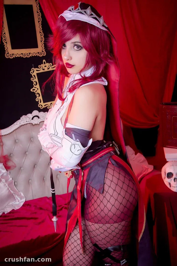 Demon Mika com um conjunto de cosplay vermelho e branco ao lado de um sofá branco capitonê contra cortinas vermelhas.