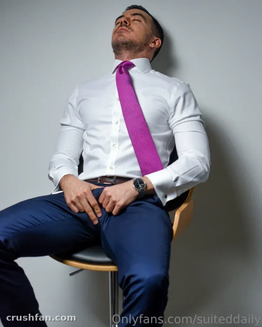 Man in a white shirt and magenta tie seated on a chair against a grey wall