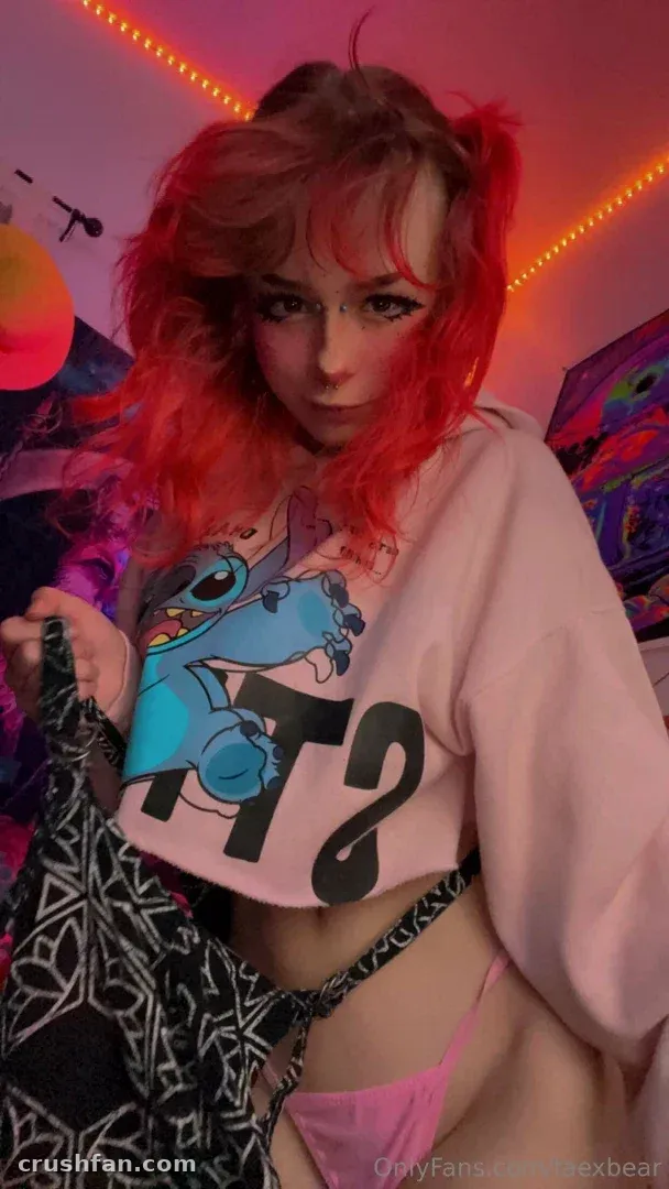 Fae with red hair in a neon-lit room wearing a pink graphic top and holding patterned clothing.