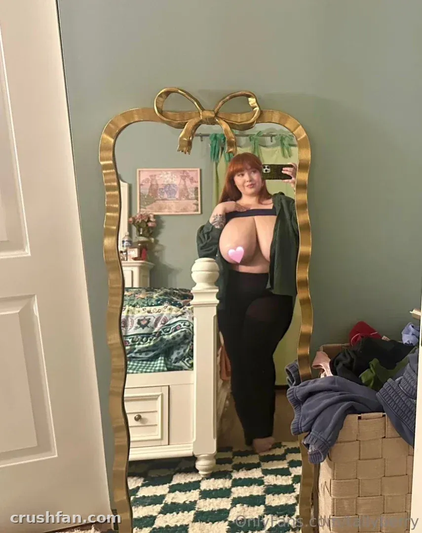 Tally com uma blusa verde aberta e legging preta tirando uma selfie no espelho em um quarto verde.