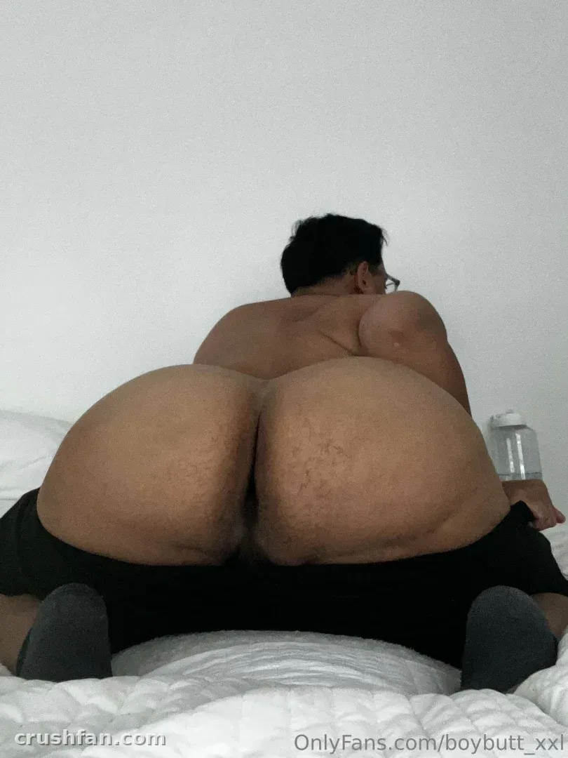 Creator BoyButtXXL liegt auf einem weißen Bett in dunkler Shorts und grauen Socken, von hinten gesehen.