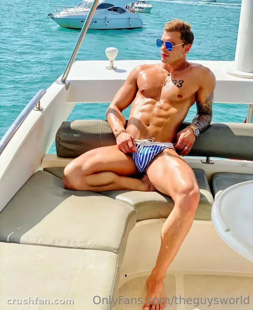 Jaky 22cm Free Page sitzt mit gekreuzten Beinen auf einem Boot in blau gestreifter Badehose und Sonnenbrille