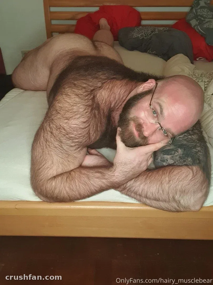 Hombre barbudo con gafas recostado en una cama con almohadas rojas y grises