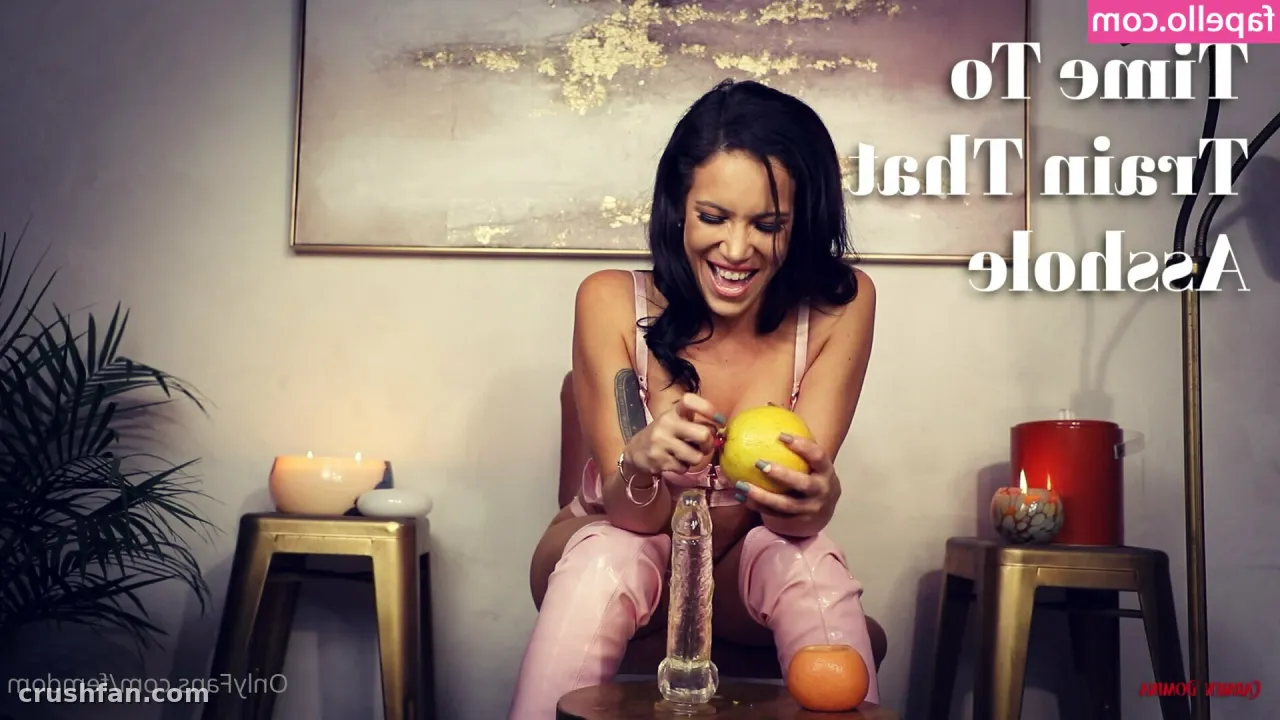 Woman in pink outfit seated indoors holding a yellow citrus fruit beside a glass decanter
