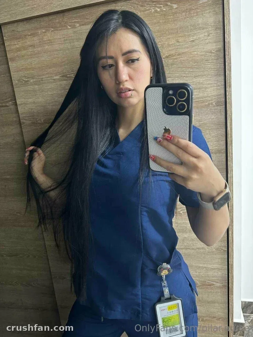 L'infirmière Pilar en tenue bleue prenant un selfie dans le miroir contre un mur lambrissé de bois.