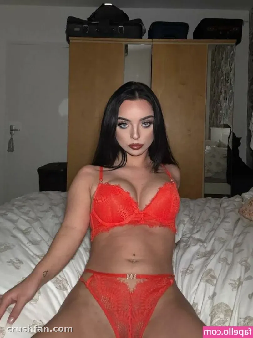 Demi in red lace lingerie seated on a white bed in a bedroom