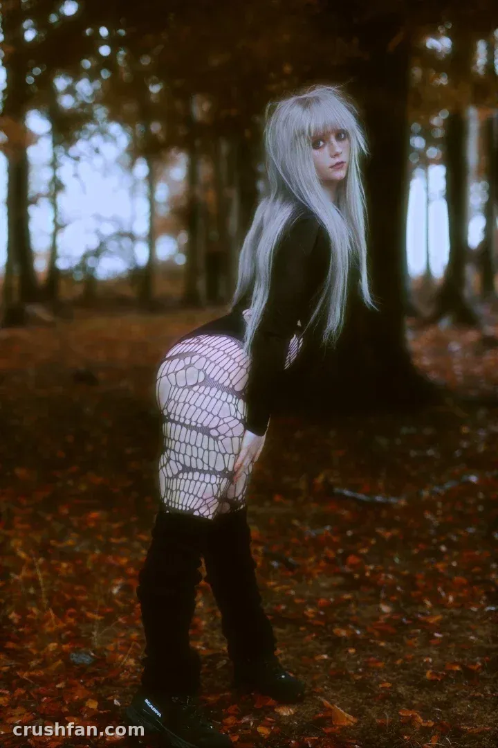 Créateur aux longs cheveux argentés posant dans une forêt d'automne vêtu de vêtements noirs et de collants à motifs.