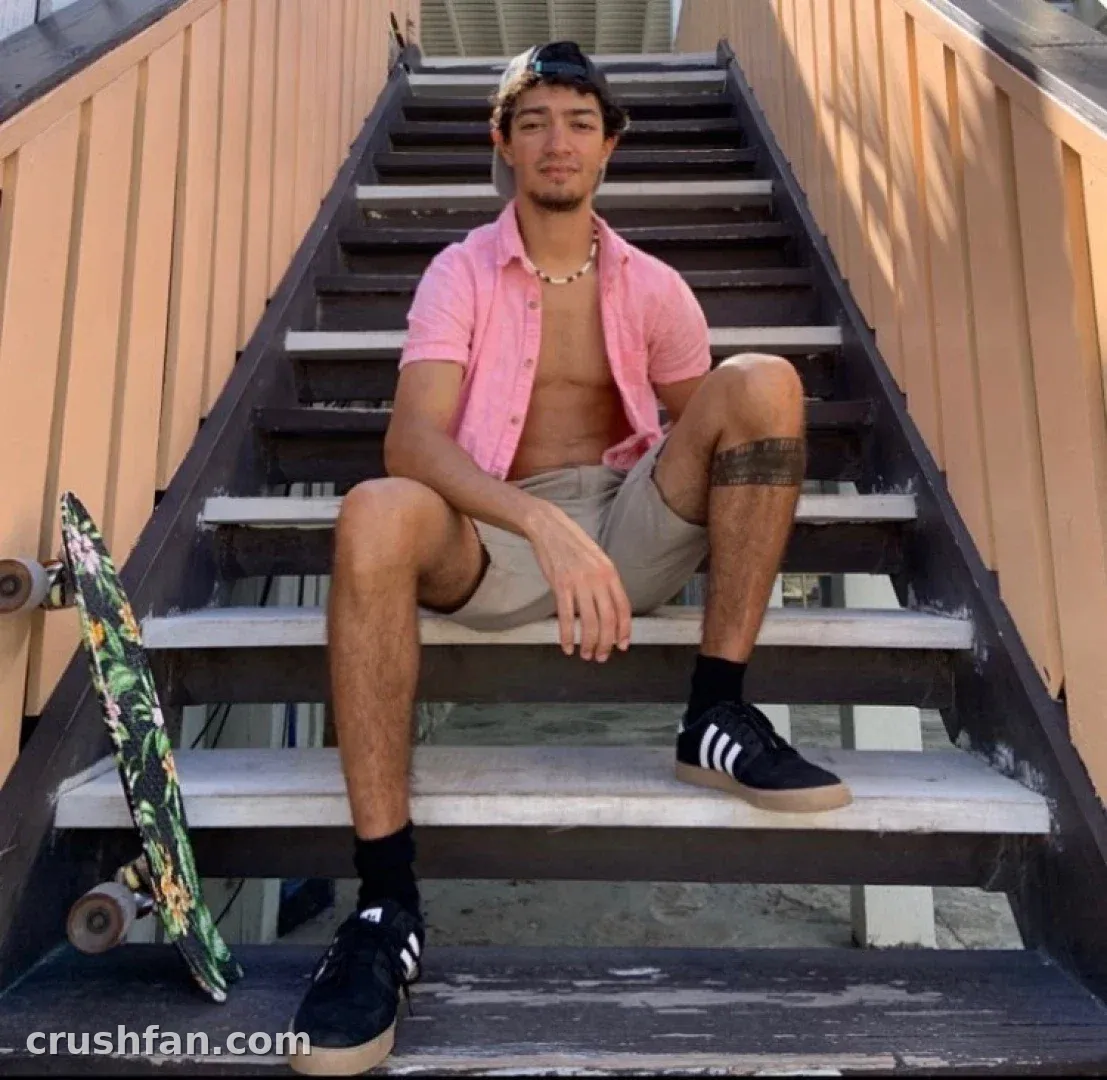Nathan Broski sentado en unas escaleras exteriores junto a un monopatín estampado con ropa informal de verano