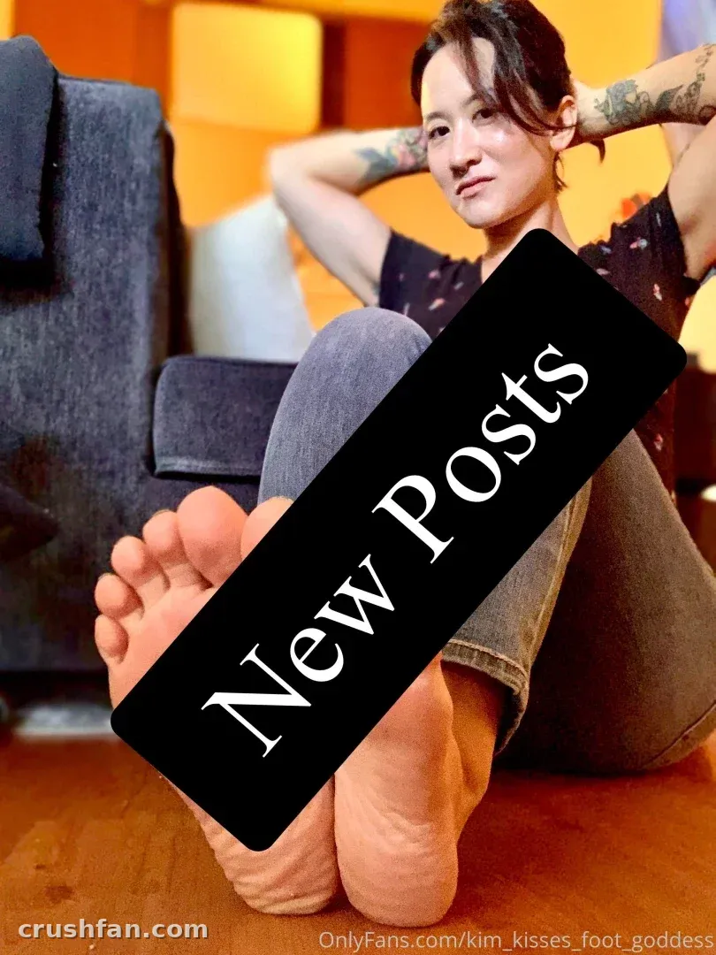 Kim Kisses sentada en un suelo de madera con los pies descalzos extendidos hacia adelante bajo una cálida iluminación interior
