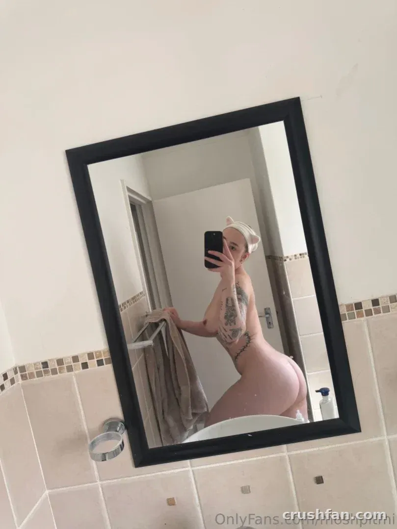 Miya macht ein Spiegel-Selfie in einem gefliesten Badezimmer und trägt einen Kopfwickel und ein rosa Outfit.