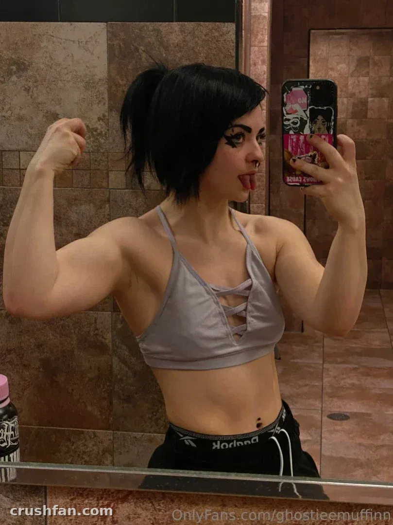Ghostiee macht ein Spiegel-Selfie in einem Fitnessstudio, flext einen Arm und streckt die Zunge heraus.