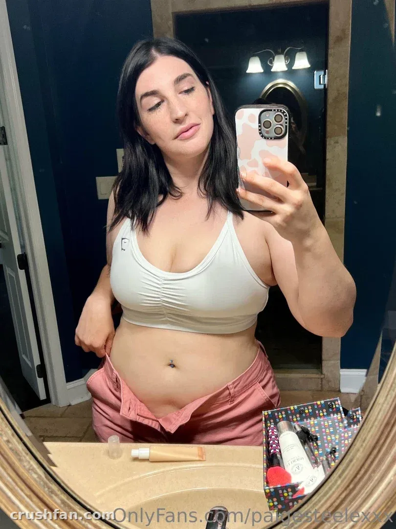 Paige Steele con un top corto blanco y pantalones rosas tomándose una selfie en el espejo en interiores
