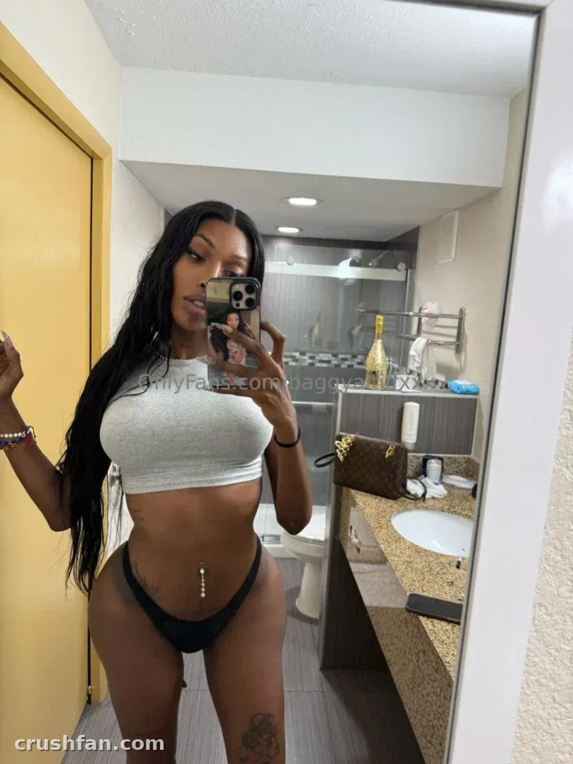 mAmi-Tiff tomándose un selfie en el espejo en un baño luminoso con un top corto gris y bragas de bikini oscuras.