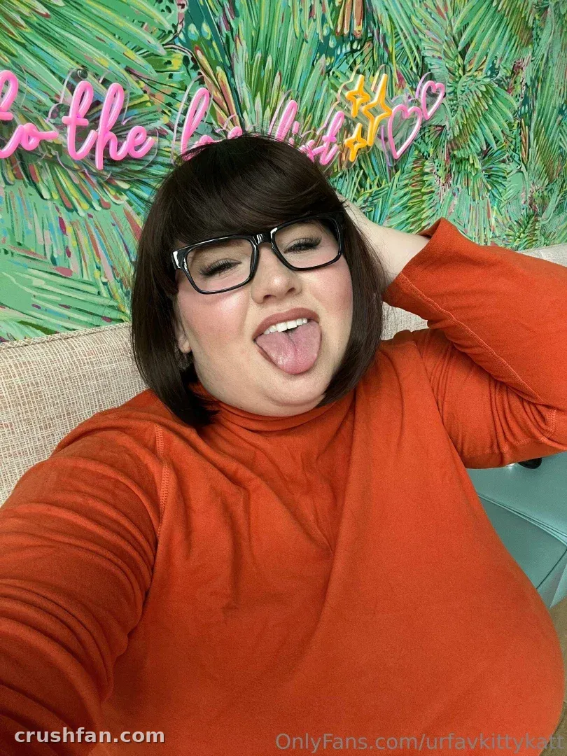 Criadora com um top laranja fazendo uma careta brincalhona na frente de uma parede verde tropical.