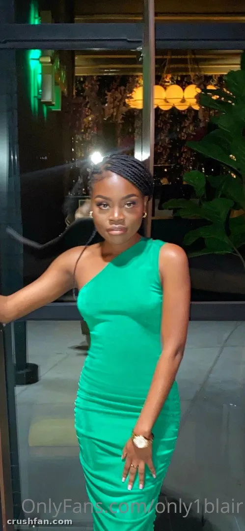 Blair in a green one-shoulder dress standing by a glass door at night