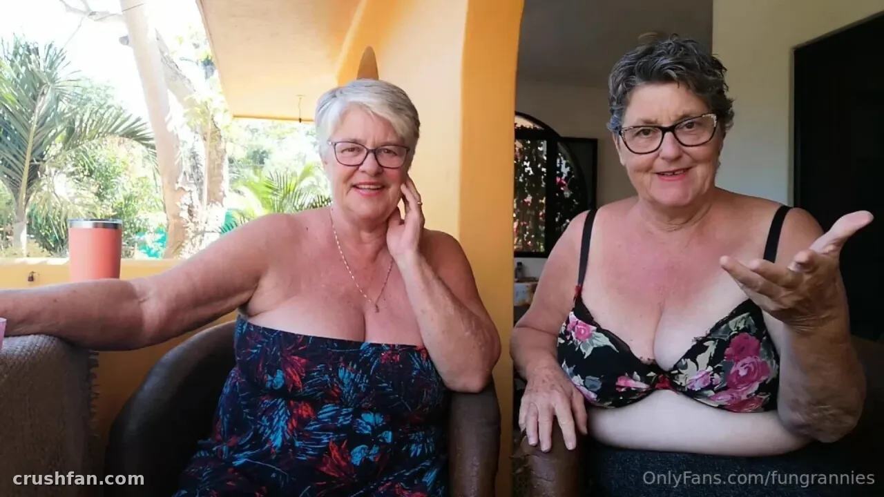 Two women with glasses on a sunny patio, one in a floral top and one in a floral bra, smiling at the camera.
