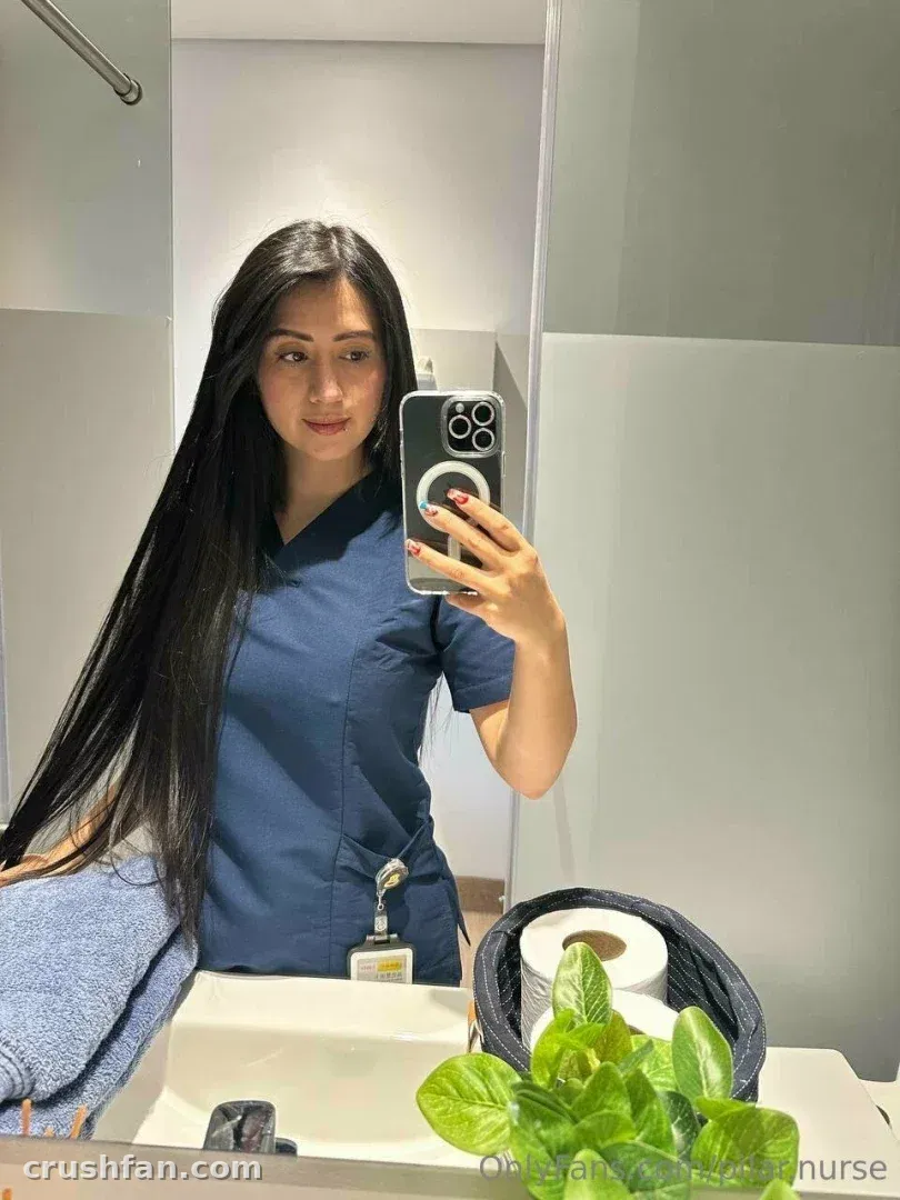 Медсестра Пилар в синей медицинской одежде делает селфи в зеркале в светлой ванной.