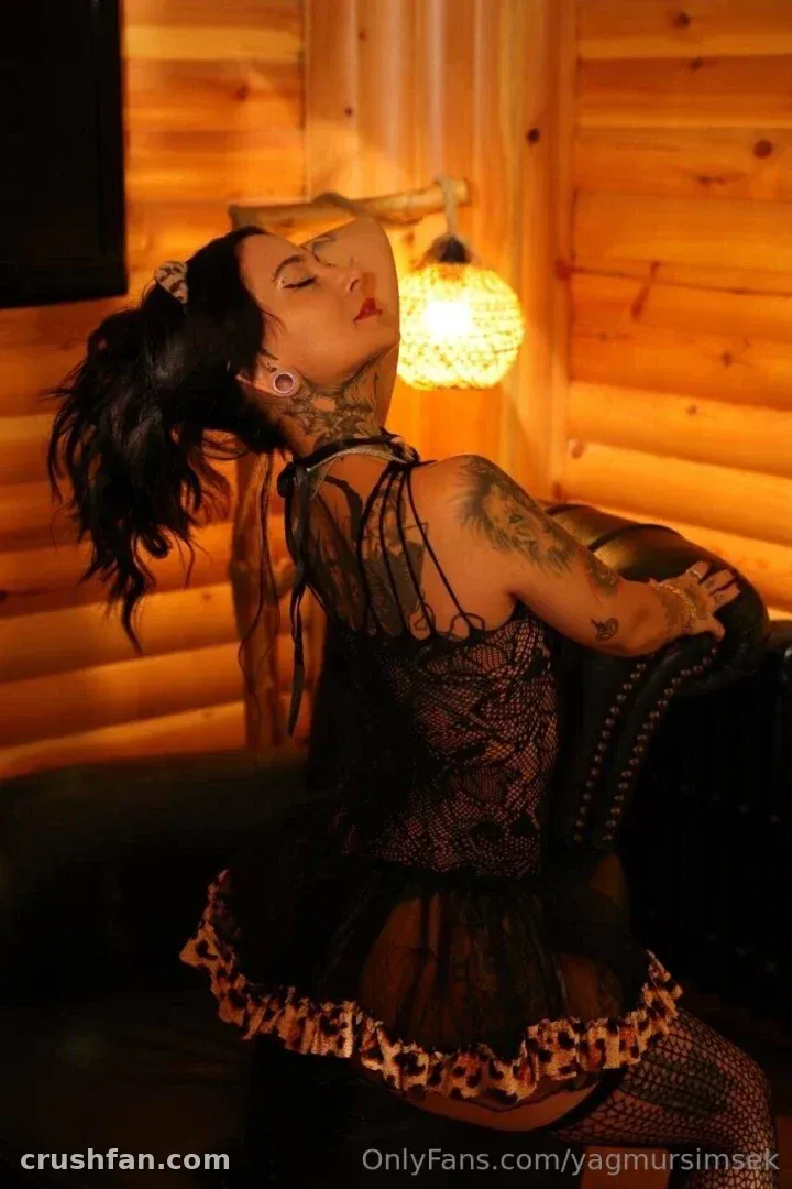 Yağmur Şimşek in a sheer top and ruffled skirt posing in a warmly lit wooden cabin.