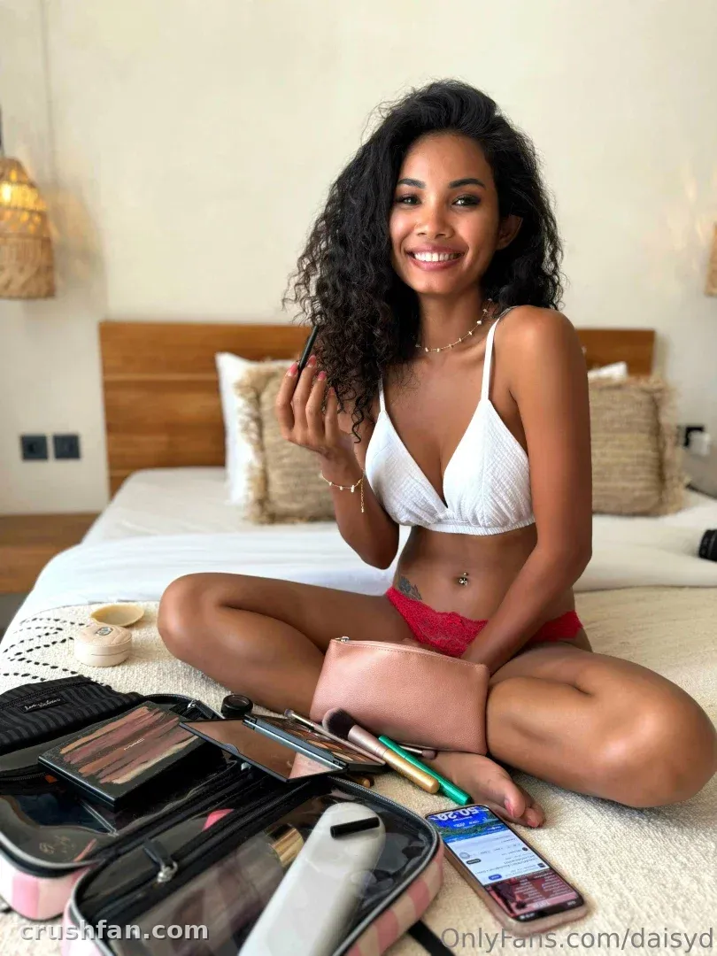 Daisyd assise sur un lit avec des produits de maquillage, portant un haut de bikini blanc et une culotte rose.