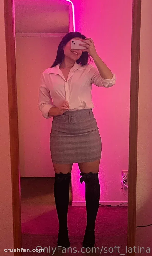Ingrid Gonzalez tomándose una selfie en el espejo con una camisa blanca y una falda gris bajo una luz rosa