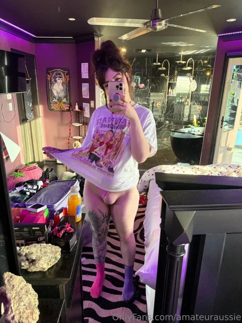 Aus Sarah prend un selfie devant le miroir dans une pièce éclairée en violet tout en relevant son t-shirt à motif.