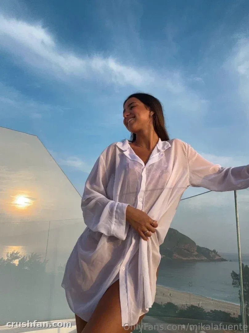 Mika in a loose white shirt on a balcony, smiling beside a sunset sea view