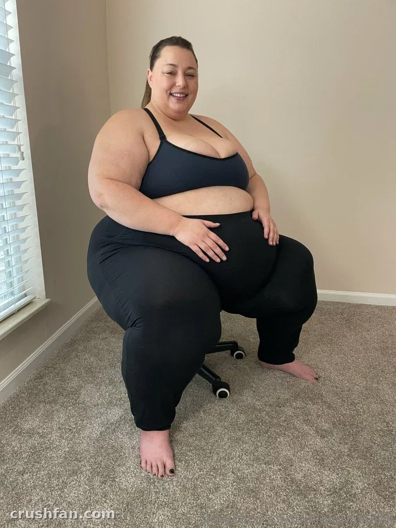 LisaLou sitzt auf einem Rollhocker und trägt einen schwarzen Sport-BH und Leggings in einem mit Teppich ausgelegten Raum.