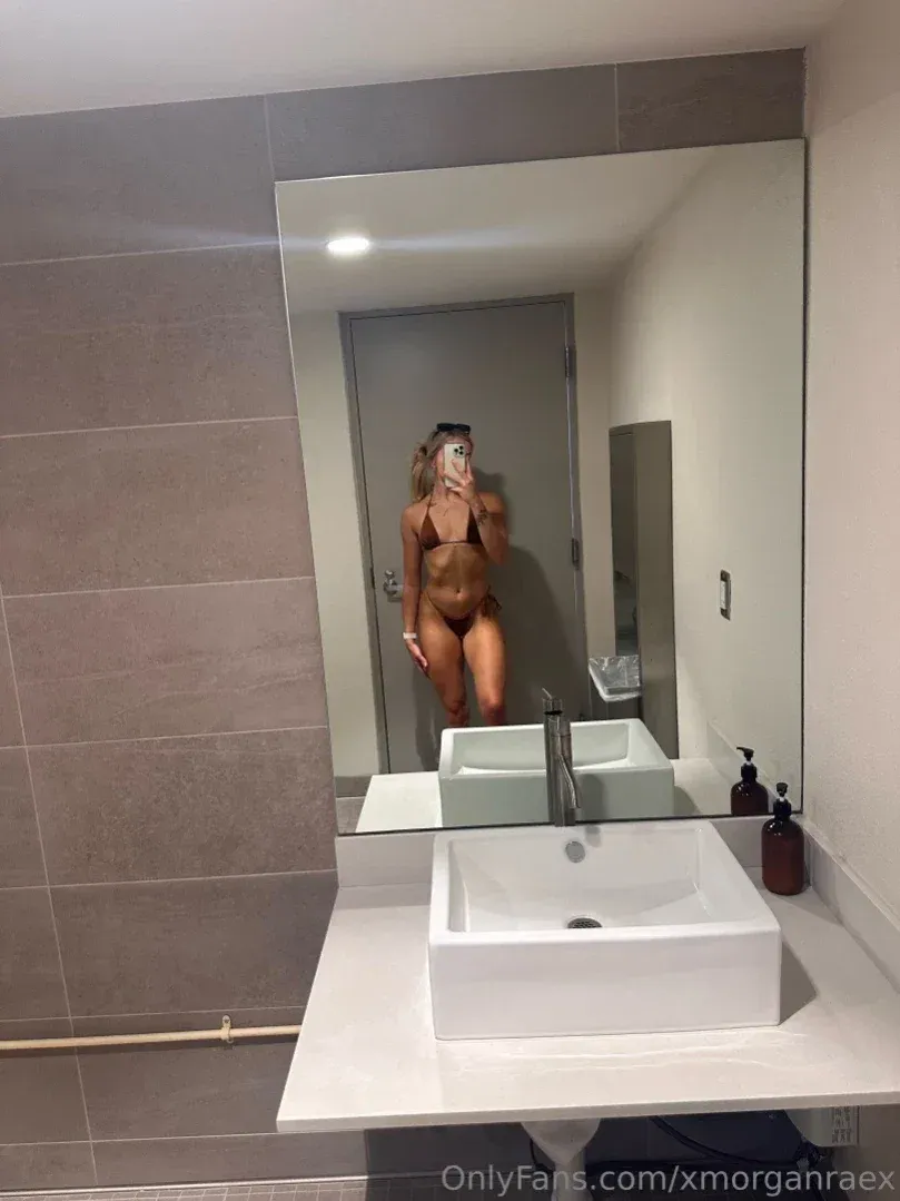 Morgan Rae prenant un selfie miroir dans une salle de bain moderne avec des carreaux gris et un lavabo blanc.